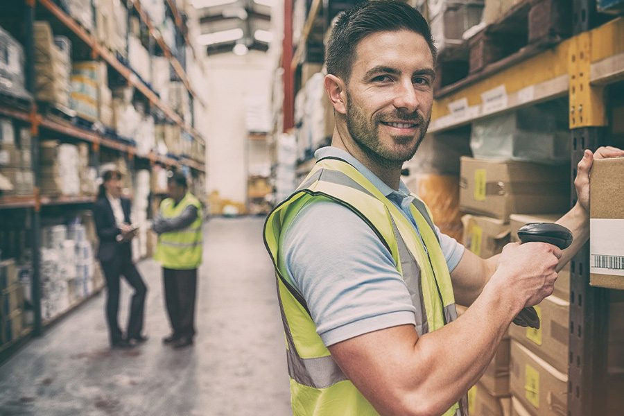 Warehousing and Logistics Insurance Portrait of a Warehouse Worker Scanning a Box While Smiling With Fellow Colleagues Blurred in the Distance